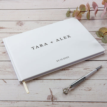 Custom wedding guest book in white which has a thick cover with a page marker. The sample has a ball point next to it and has couple's name as "TARA + ALEX" and the wedding date as "23.10.2023"